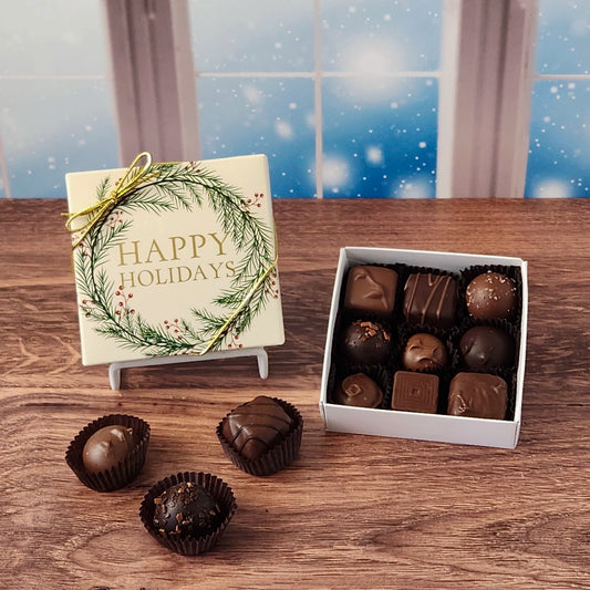 9 piece box of chocolates with decorative Happy Holidays wreath cover on a wooden surface and snowy background.