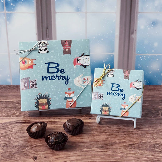 Two decorative boxes with animal illustrations and 'Be merry' text on a wooden surface with a snowy window background.
