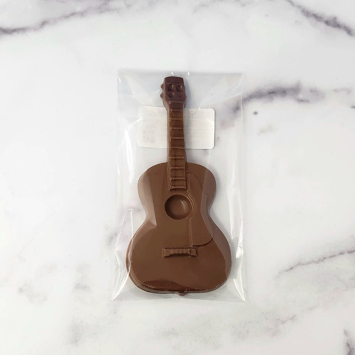 A chocolate guitar-shaped item sealed in a clear plastic bag, placed on a white marble surface.