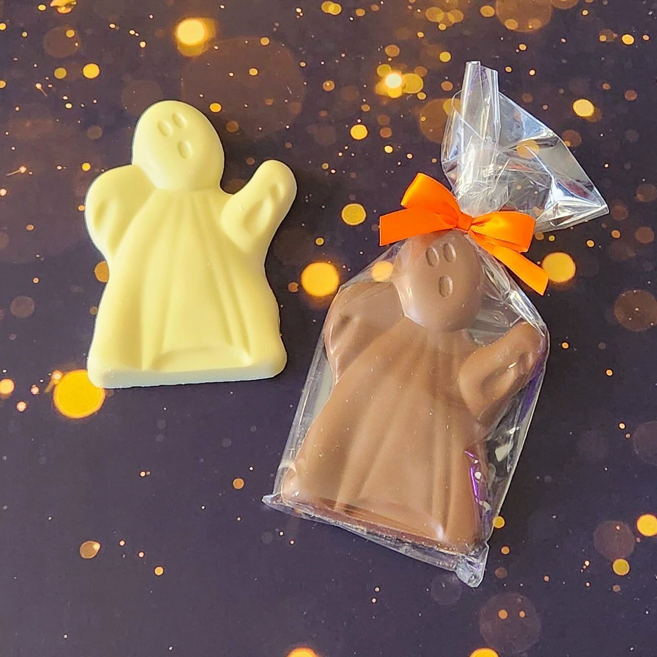 Two Haunted Chocolate Ghosts, one made of white chocolate and the other in milk chocolate. The milk chocolate ghost is packaged in a clear bag tied with an orange bow, showcasing its festive Halloween theme. The white chocolate ghost is unwrapped, highlighting the detailed ghost shape of both treats.