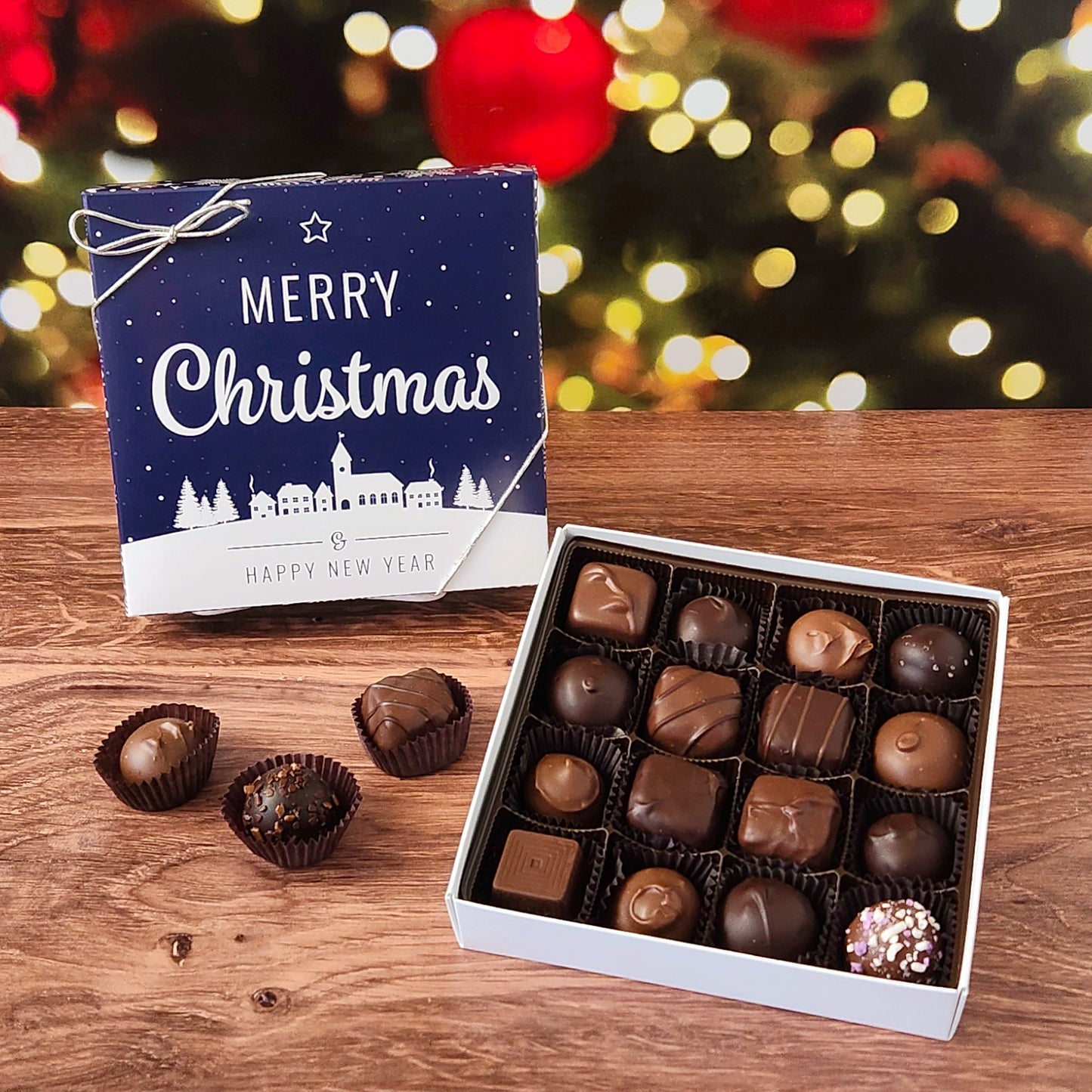 Box of chocolates with a 'Merry Christmas' box on a wooden surface with festive lights in the background.