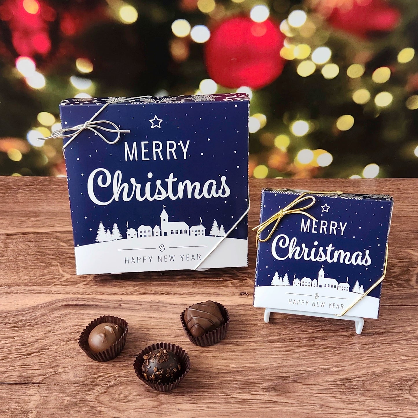 Two 'Merry Christmas' chocolate boxes on a wooden surface with blurred Christmas lights in the background.