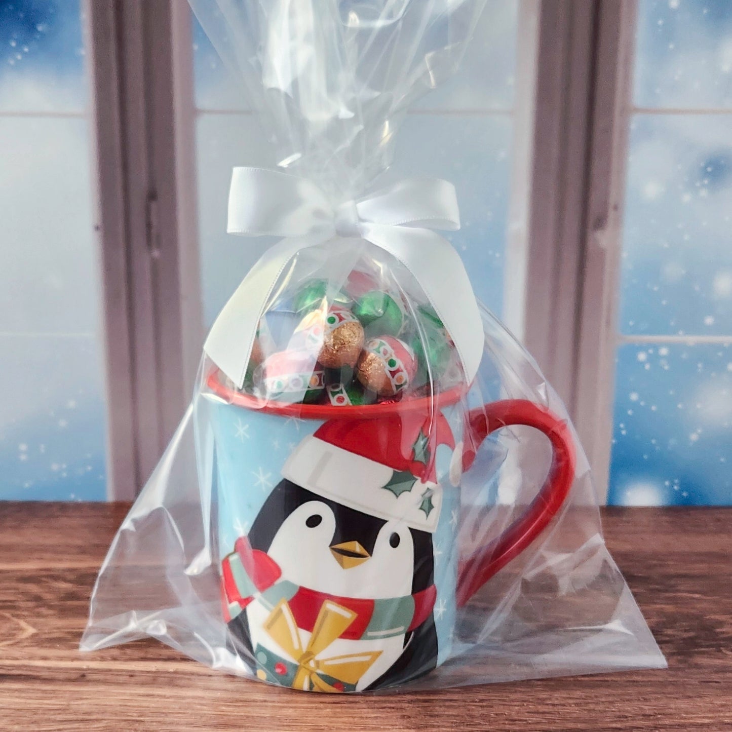 A penguin-themed ceramic mug wrapped in a clear cellophane gift bag with a white ribbon, filled with assorted chocolates, set on a wooden table in front of a snowy window background.