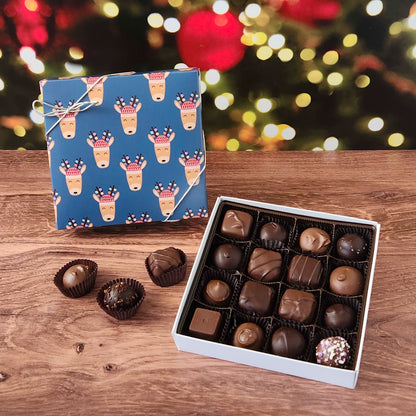 Box of chocolates with a blue reindeer-patterned box on a wooden surface with a blurred Christmas tree background.