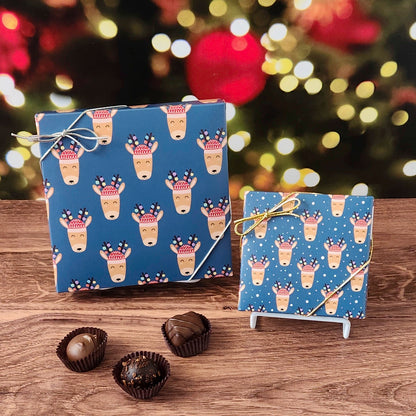 Box of chocolates with a blue reindeer-patterned box on a wooden surface with a blurred Christmas tree background.