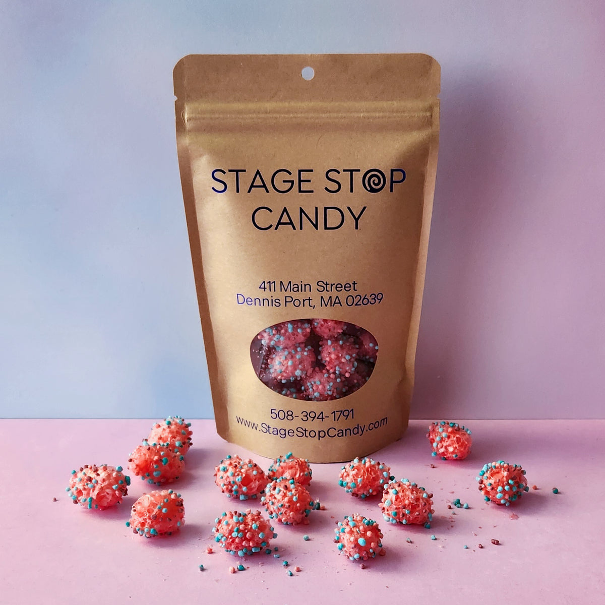 A kraft paper stand-up pouch labeled 'STAGE STOP CANDY' with a clear window showing pink and red sprinkled candy inside, with several loose candy pieces scattered in front on a pink surface against a pastel blue-to-pink gradient background.