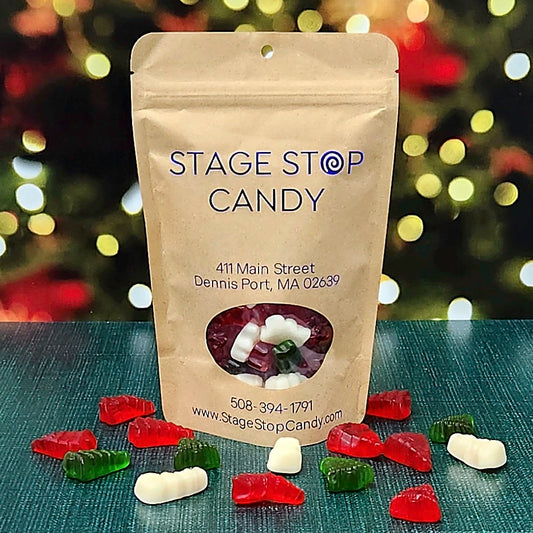 Gummi Snowmen and Trees in a kraft bag, with red and green tree gummis and white snowmen scattered on a teal table before holiday lights