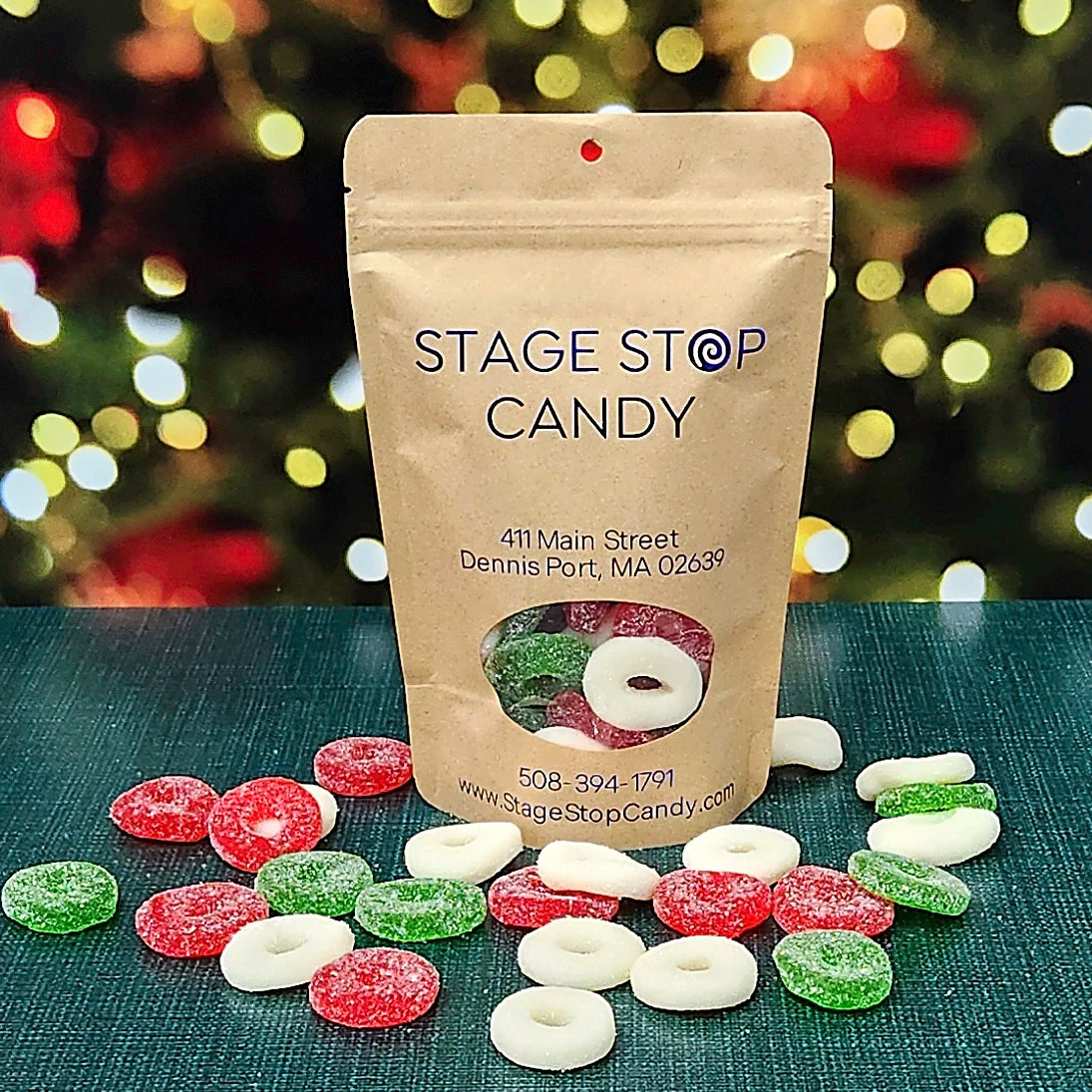 Gummi Wreaths in a kraft bag, with sugar-sanded red, green and white rings spread on a teal table before holiday lights