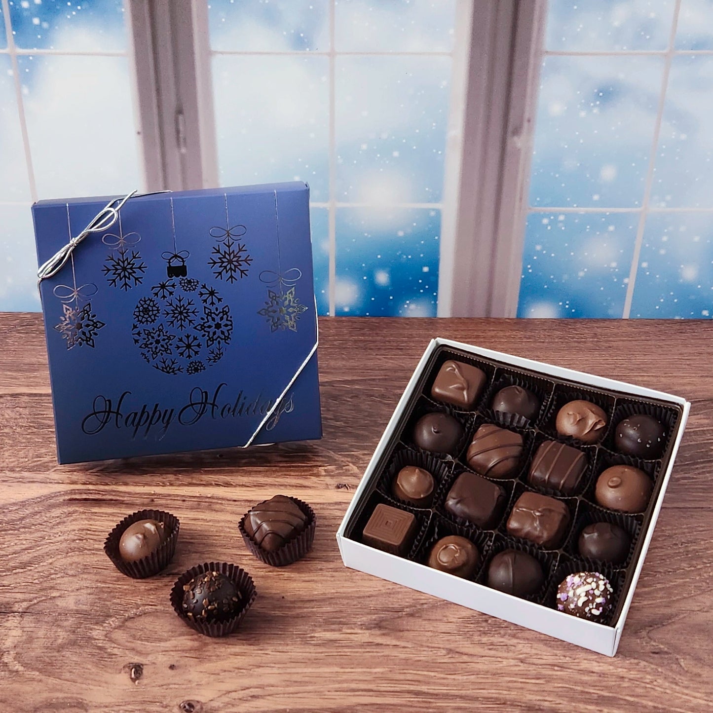 16 piece box of chocolates with  decorative Happy Holidays ornament cover on a wooden surface and snowy background.