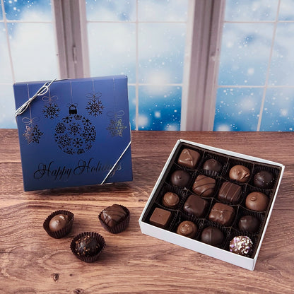 16 piece box of chocolates with  decorative Happy Holidays ornament cover on a wooden surface and snowy background.