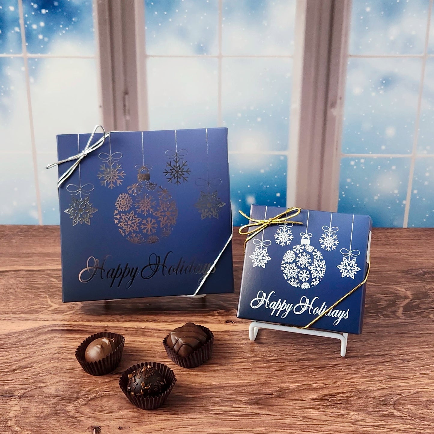 16 piece box of chocolates with  decorative Happy Holidays ornament cover on a wooden surface and snowy background.