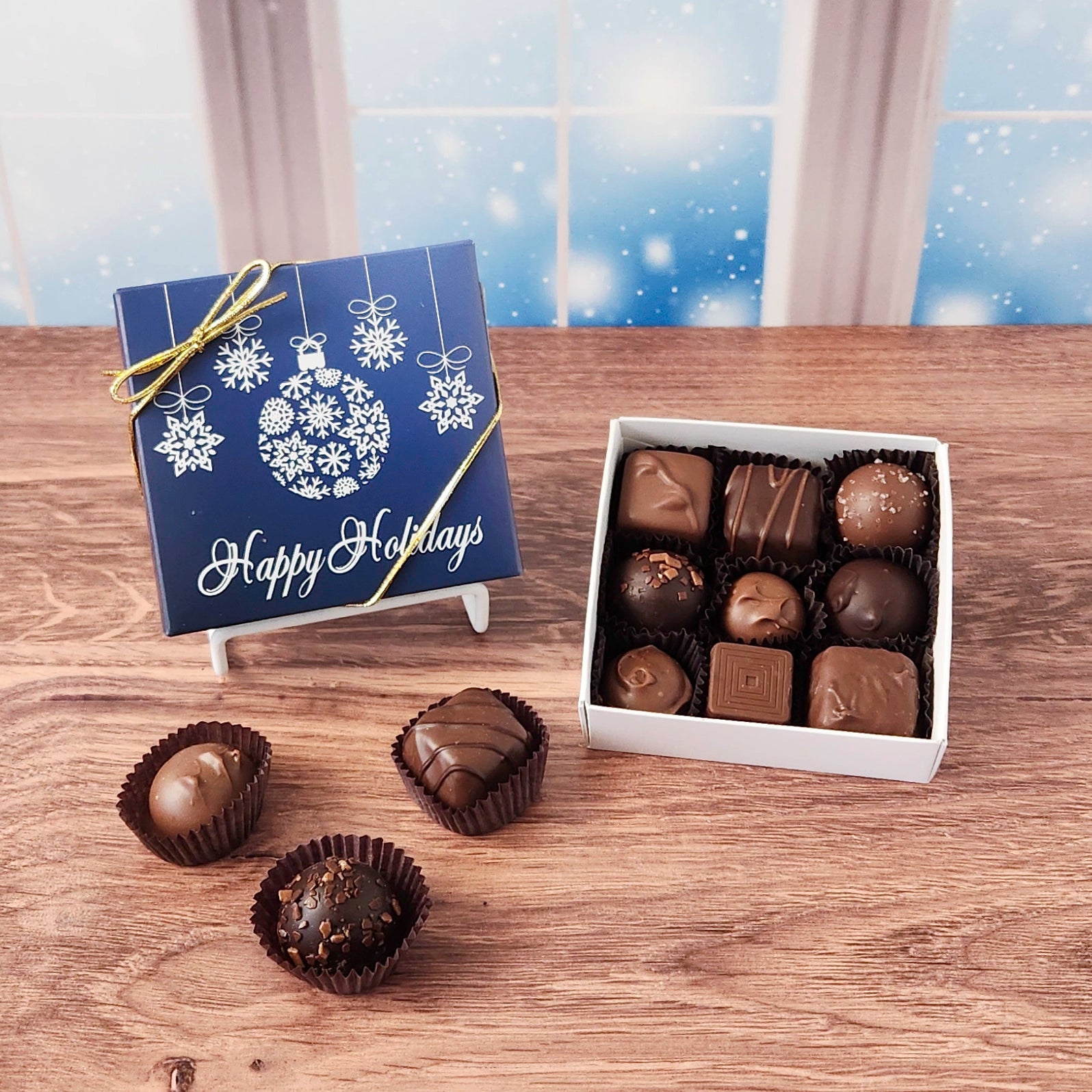 9 piece box of chocolates with  decorative Happy Holidays ornament cover on a wooden surface and snowy background.