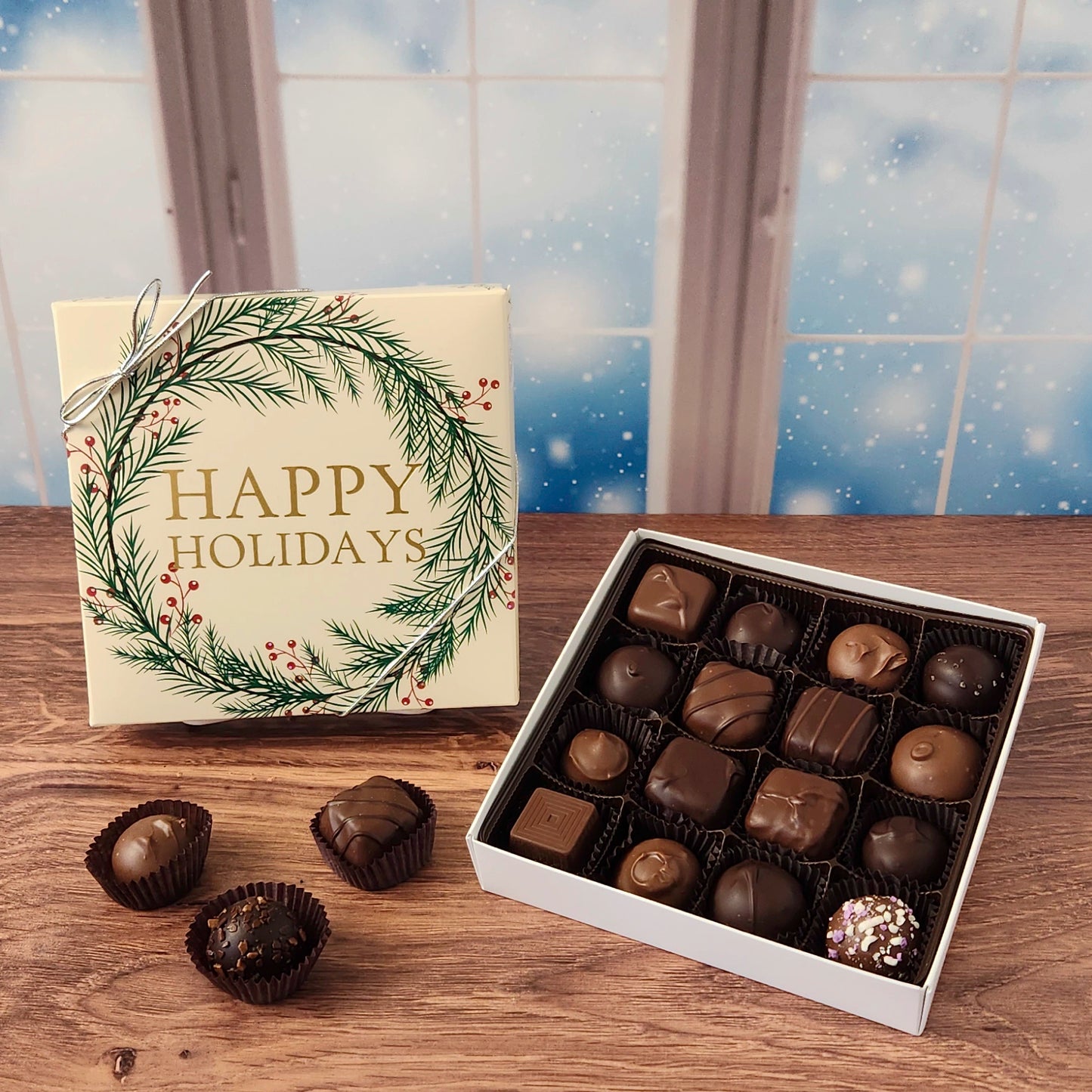 16 piece box of chocolates with  decorative Happy Holidays wreath cover on a wooden surface and snowy background.