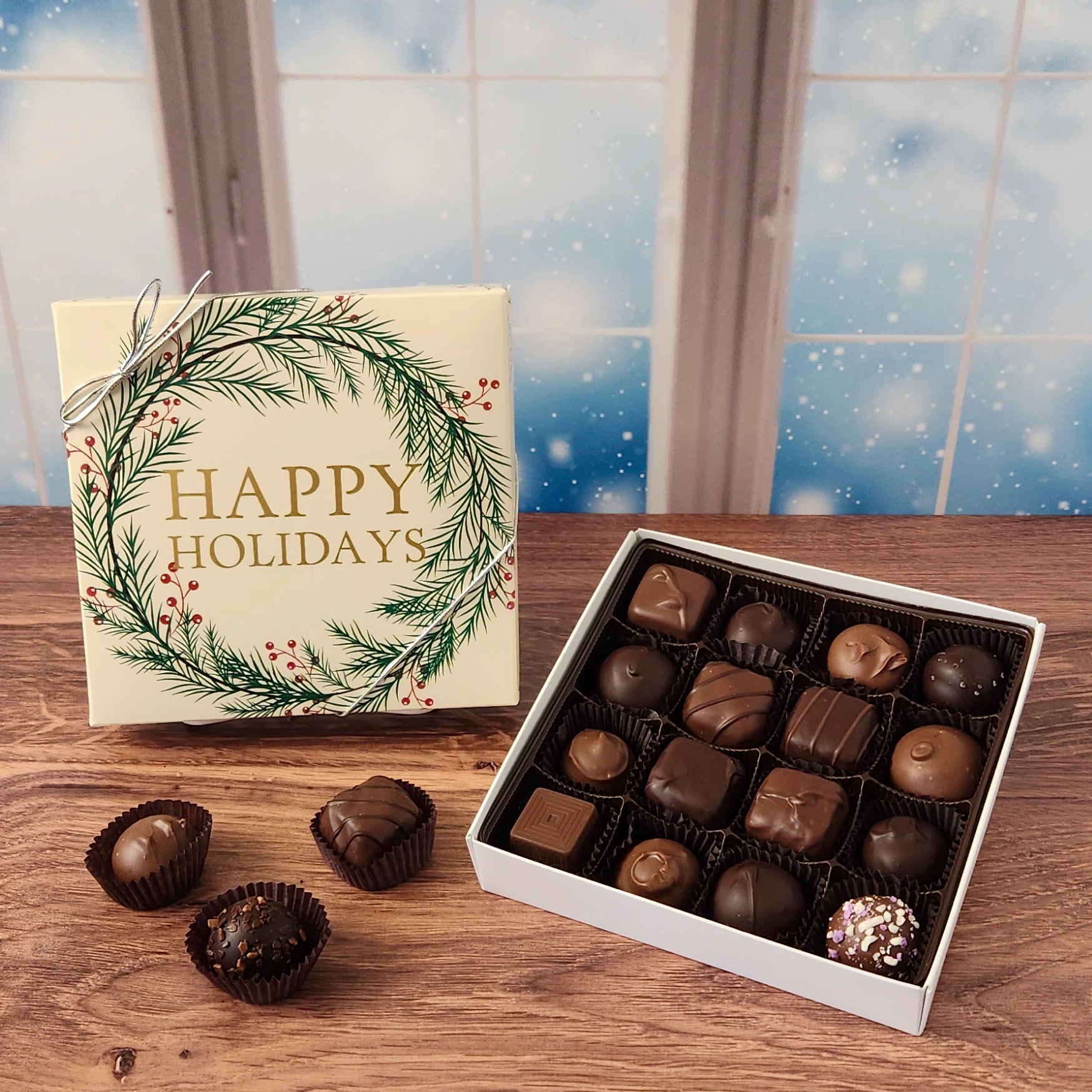 16 piece box of chocolates with  decorative Happy Holidays wreath cover on a wooden surface and snowy background.