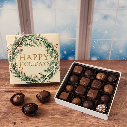 16 piece box of chocolates with  decorative Happy Holidays wreath cover on a wooden surface and snowy background.