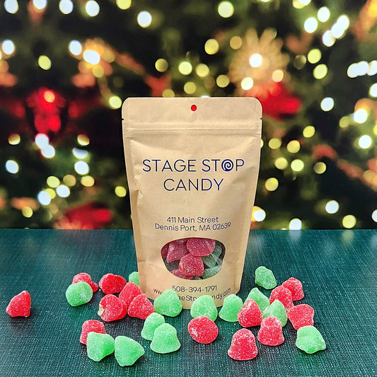 Jingle Bell Jells in a kraft bag, with sugar-coated red and green bell shapes scattered on a teal table before holiday lights