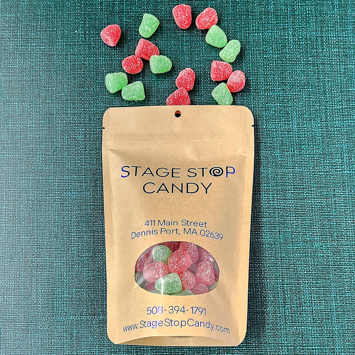 A kraft paper stand-up pouch labeled 'STAGE STOP CANDY' with a clear window showing assorted gummy candies inside, plus several loose spice drop gummies scattered above it on a textured teal background.
