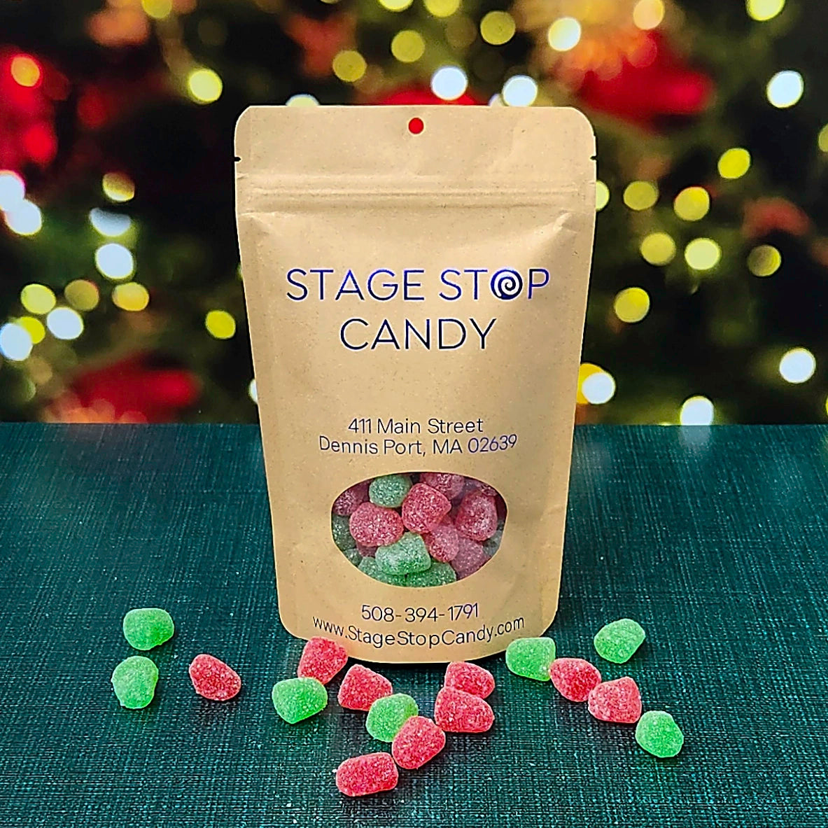 A kraft paper stand-up pouch labeled 'STAGE STOP CANDY' with a clear window showing pink and green sugar-coated candies inside, set on a dark teal surface with scattered matching candies in front and a blurred festive tree of lights in the background.