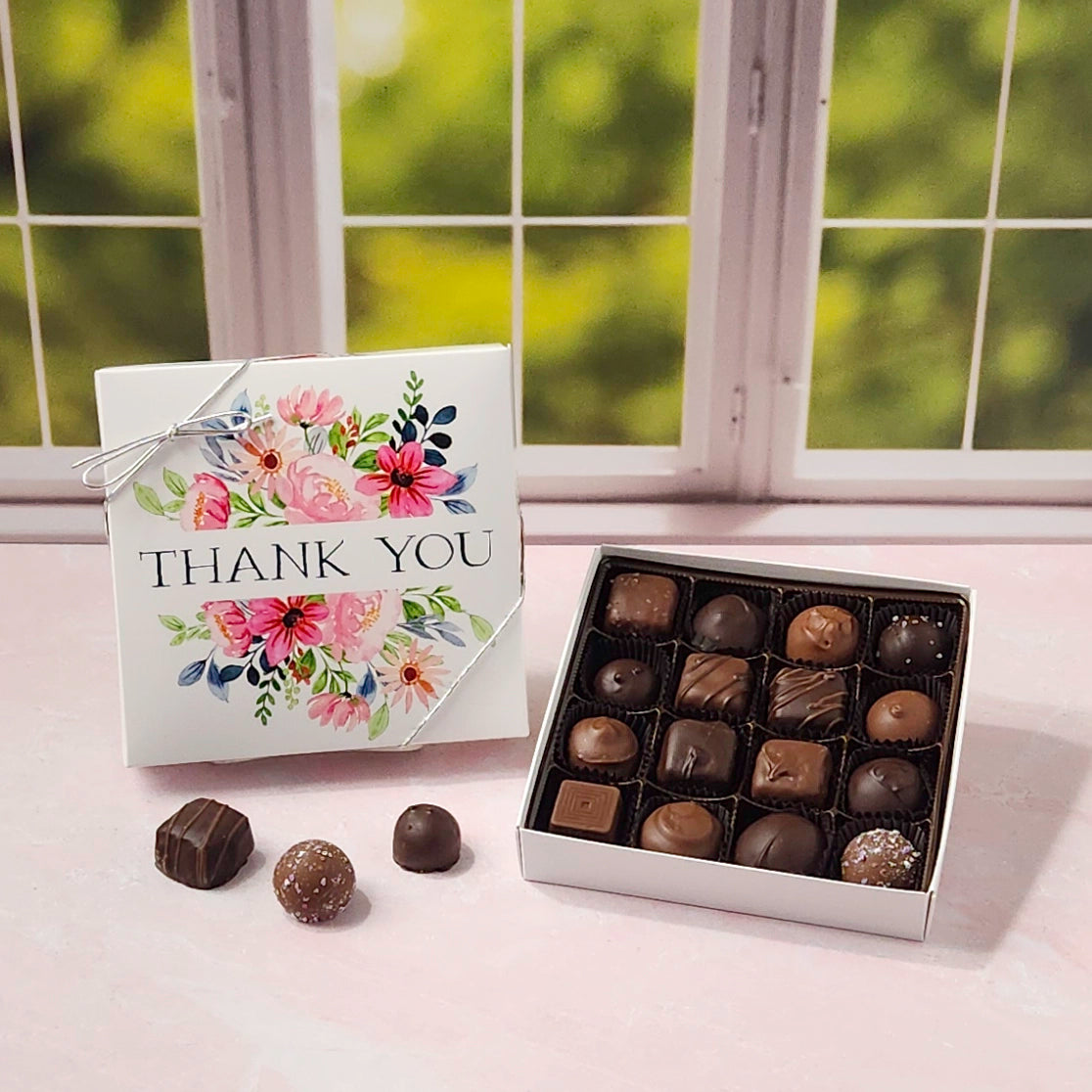 Box of chocolates with a 'Thank You' box on a windowsill with greenery outside.