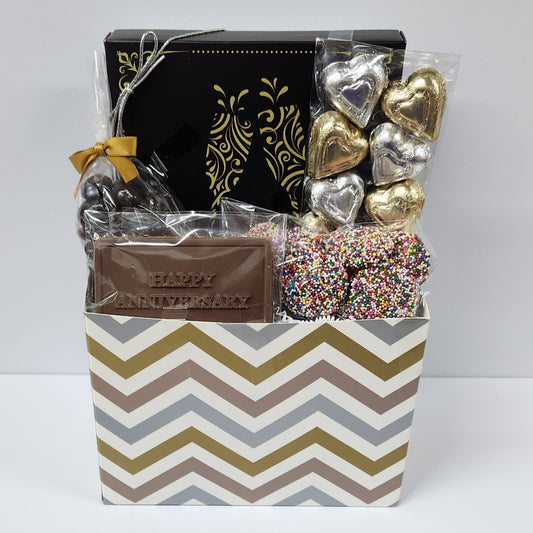 Happy Anniversary Chocolate Gift Basket features silver and gold foiled milk chocolate hearts, dark chocolate-covered cranberries, milk chocolate nonpareils, a milk chocolate card that says "Happy Anniversary", and a 16 piece gift box containing creams, caramels, meltaways, and truffles